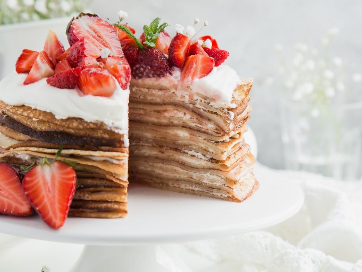 Die Pfannkuchen-Torte auf einer weißen Tortenplatte vor hellem Hintergrund und auf einem weißen Tisch. Alles von der Seite fotografiert.