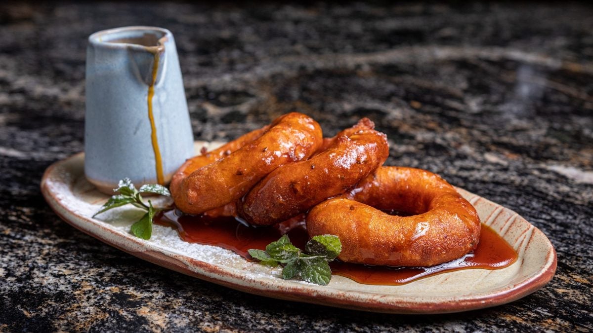 Drei Picarones auf einem Serviertablett mit Sirup und einer Karaffe daneben.