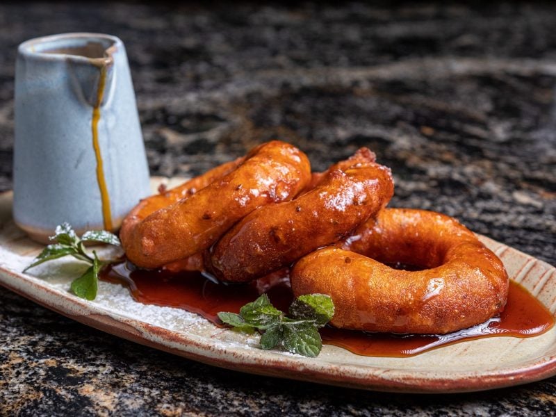 Drei Picarones auf einem Serviertablett mit Sirup und einer Karaffe daneben.