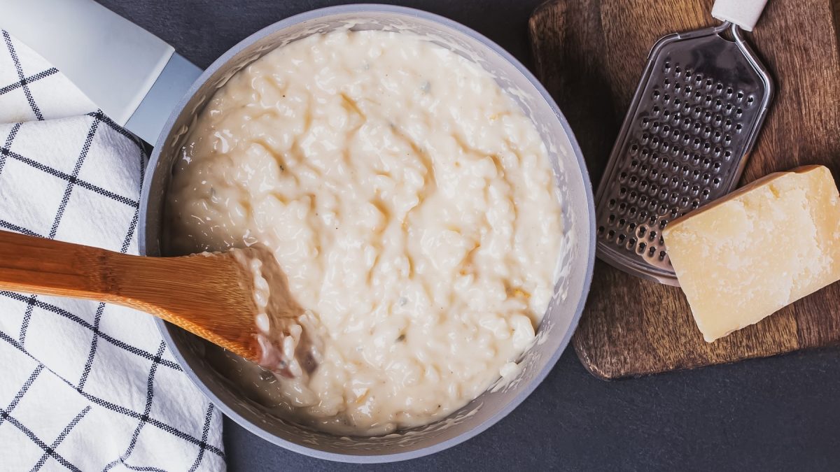 Cremiges Risotto in einem Topf mit einem Holzlöffel gerührt, daneben ein Stück Parmesan und eine Reibe auf einem Holzbrett.