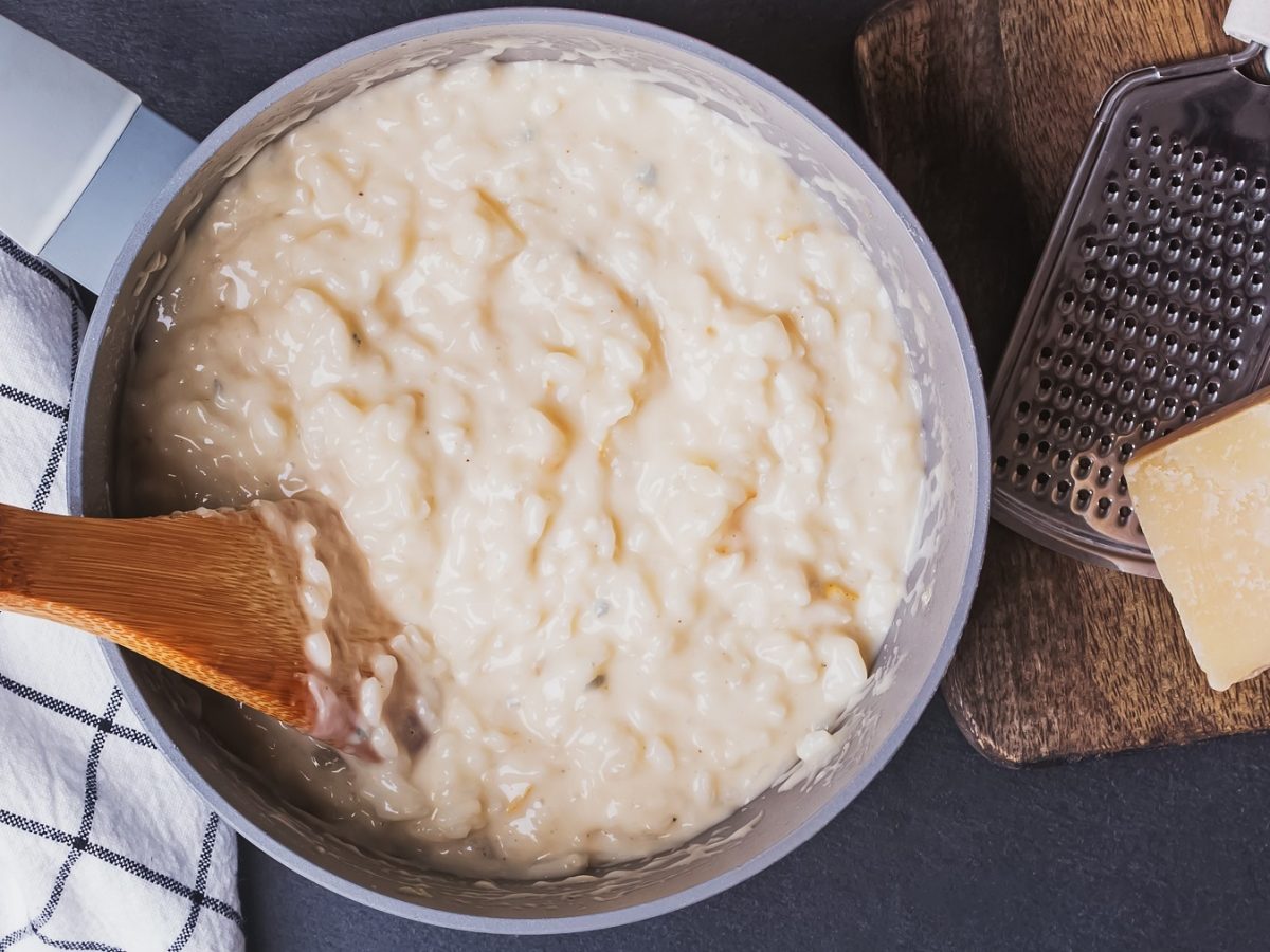 Cremiges Risotto in einem Topf mit einem Holzlöffel gerührt, daneben ein Stück Parmesan und eine Reibe auf einem Holzbrett.