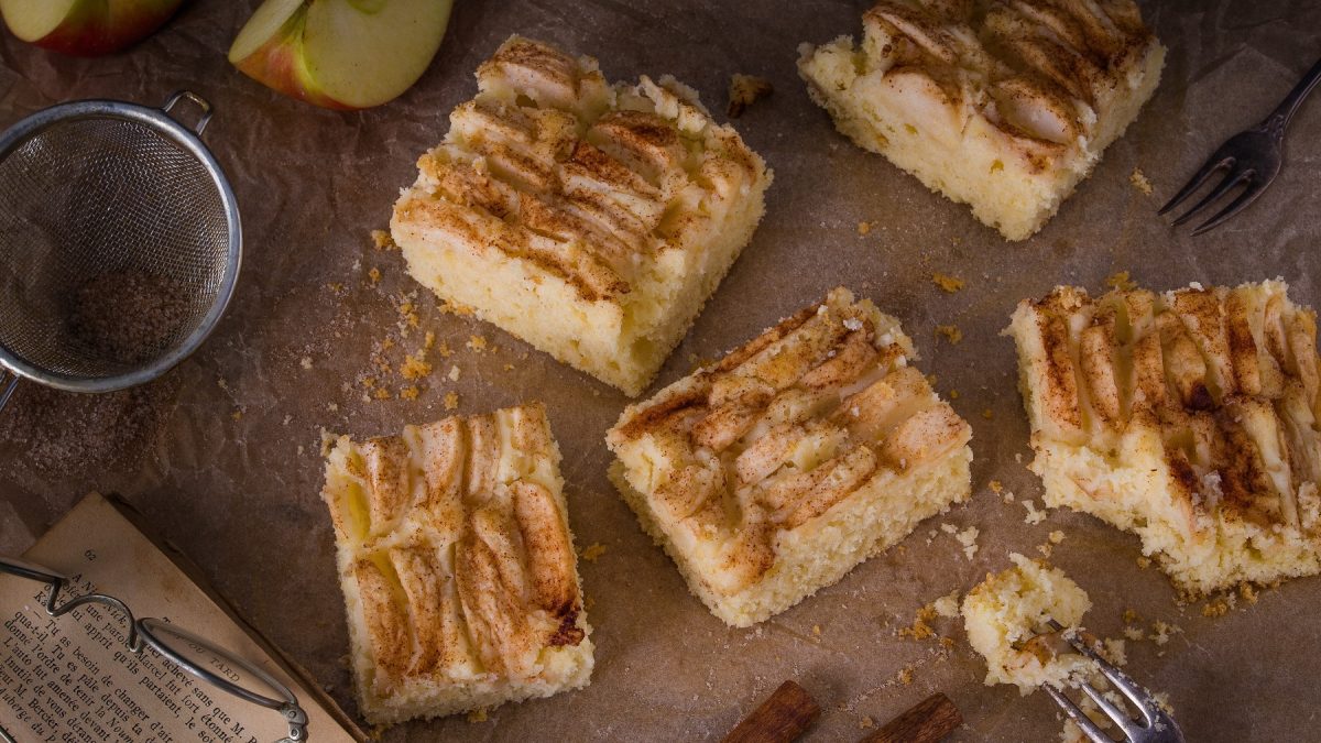 Stücke vom schwedischen Apfelkuchen vom Blech auf Backpapier, daneben Sieb und Apfelschnitze.