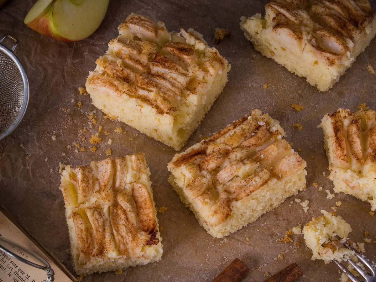 Stücke vom schwedischen Apfelkuchen vom Blech auf Backpapier, daneben Sieb und Apfelschnitze.