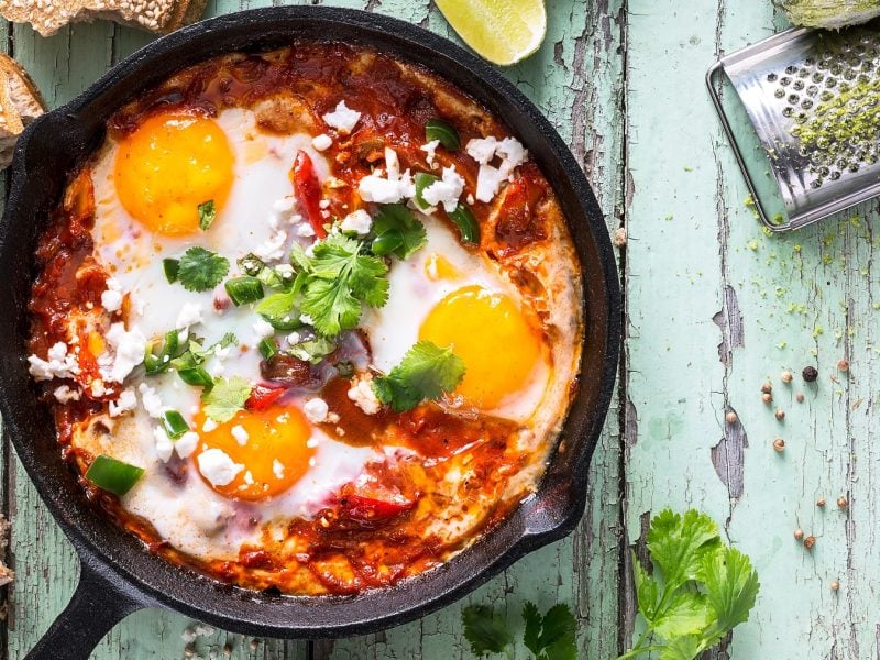 Das Shakshuka in einer gusseisernen Pfanne auf einem rustikalen Holztishc mit grünlicher Farbe direkt von oben fotografiert.