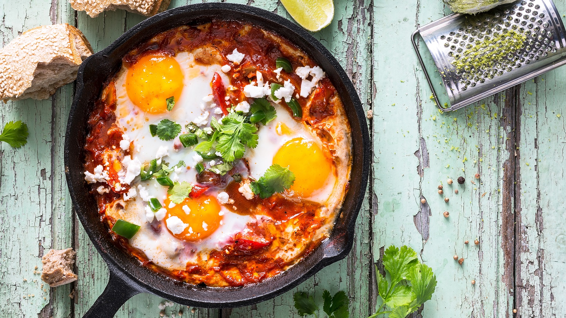 Das Shakshuka in einer gusseisernen Pfanne auf einem rustikalen Holztishc mit grünlicher Farbe direkt von oben fotografiert.