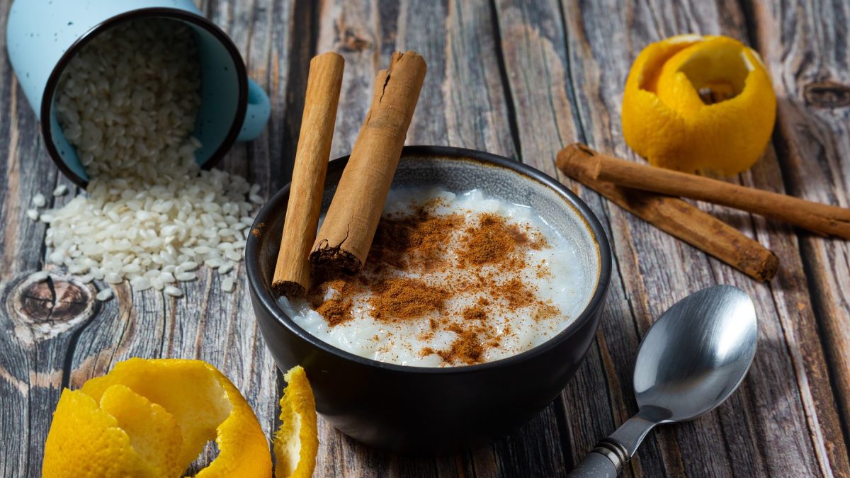 Spanischer Milchreis Arroz con leche in einer Schüssel mit Zimt, Zimtstangen. Daneben Zitronenschale, eine umgekippte Dose mit Reis und ein Löffel.