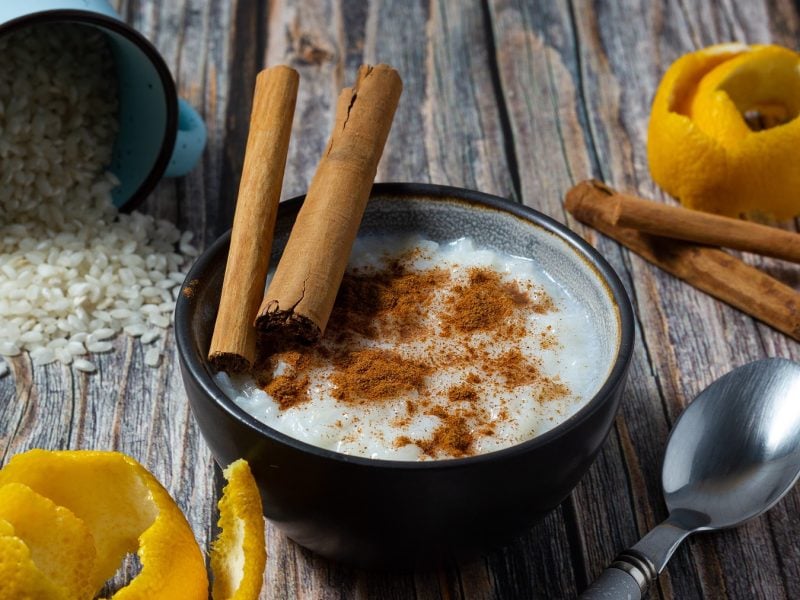 Spanischer Milchreis Arroz con leche in einer Schüssel mit Zimt, Zimtstangen. Daneben Zitronenschale, eine umgekippte Dose mit Reis und ein Löffel.