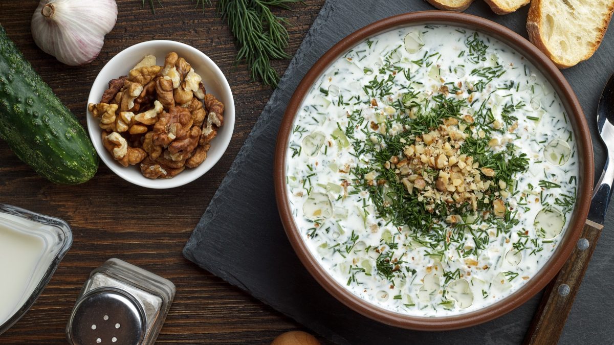 Eine Schale cremige Tarator-Suppe mit Gurkenstücken und gehacktem Dill, garniert mit gehackten Walnüssen, daneben Knoblauch, Walnüsse, Gurke und Baguettescheiben auf Holzuntergrund.