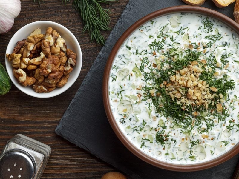 Eine Schale cremige Tarator-Suppe mit Gurkenstücken und gehacktem Dill, garniert mit gehackten Walnüssen, daneben Knoblauch, Walnüsse, Gurke und Baguettescheiben auf Holzuntergrund.