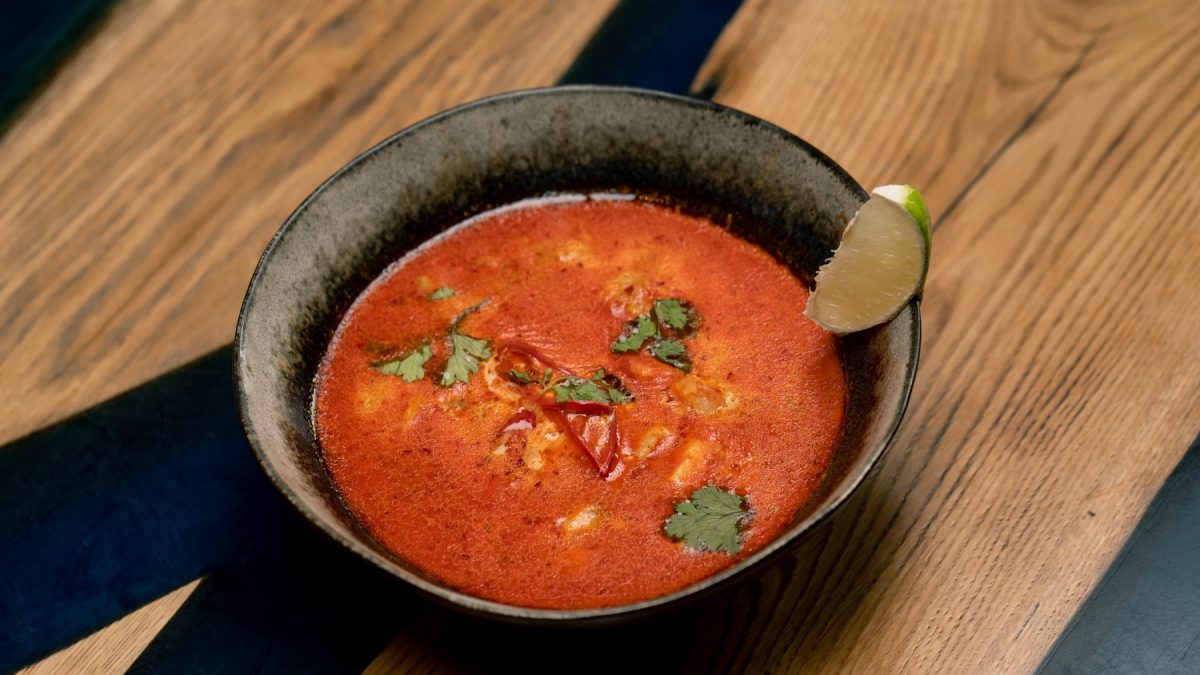 Eine dunkle Schüssel mit der Tom Yam Gai Suppe auf einem Holztisch mit schwarzen Elementen im Untergrund von schräg oben fotografiert. Am Rand des Tellers ist noch eine Limettenspalte als Garnitur.