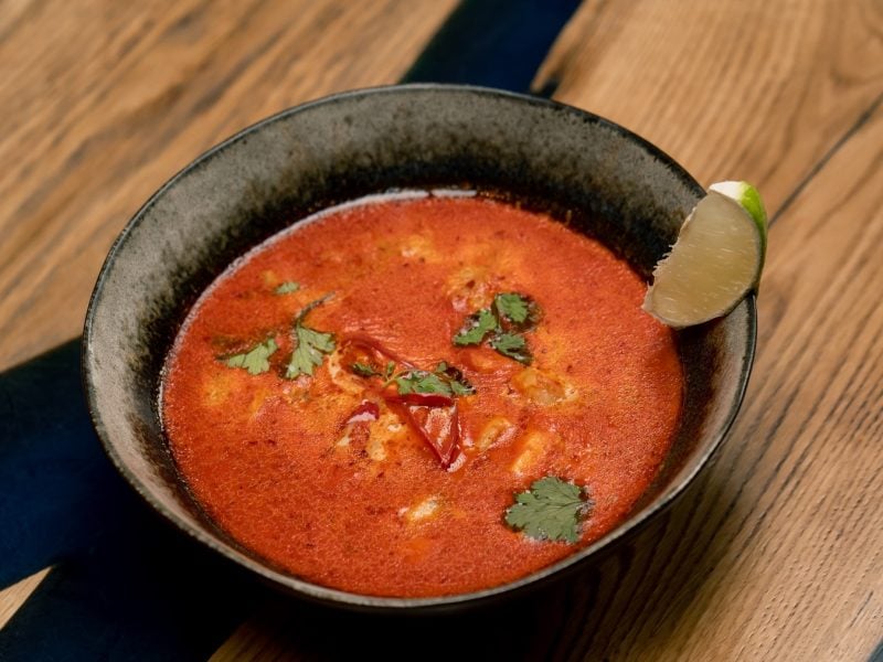 Eine dunkle Schüssel mit der Tom Yam Gai Suppe auf einem Holztisch mit schwarzen Elementen im Untergrund von schräg oben fotografiert. Am Rand des Tellers ist noch eine Limettenspalte als Garnitur.