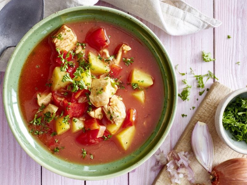 Tomatensuppe mit Huhn in einer Schüssel auf weißem Hintergrund. Links ein Geschirrtuch und ein Löffel, rechts ein Schneidebrett mit Petersilie und einer Zwiebel.