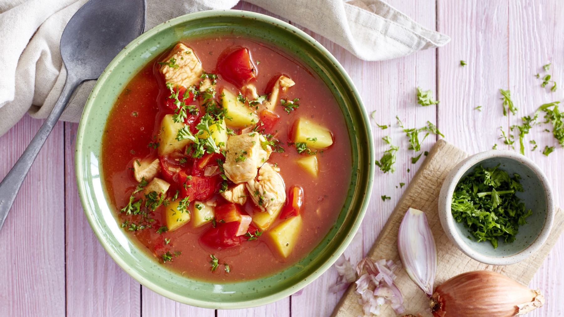 Tomatensuppe mit Huhn in einer Schüssel auf weißem Hintergrund. Links ein Geschirrtuch und ein Löffel, rechts ein Schneidebrett mit Petersilie und einer Zwiebel.