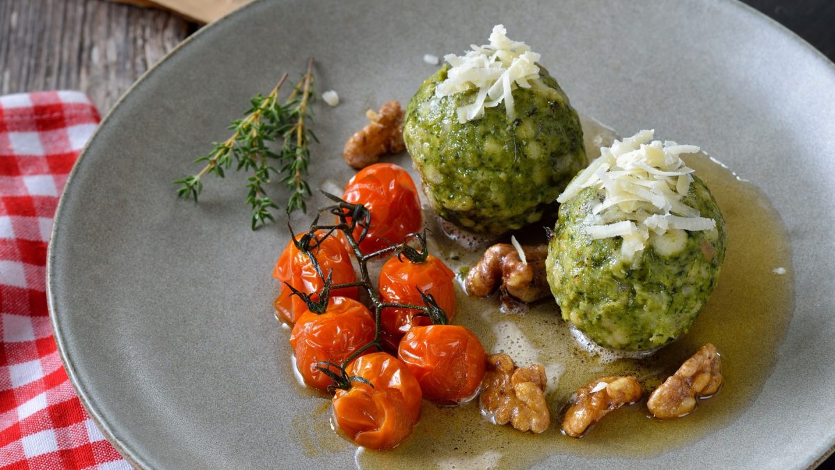 Vegane Spinatknödel mit veganer Butter und veganem Parmesan auf einem grauen Teller auf rot-weiß-kariertem Hintergrund.