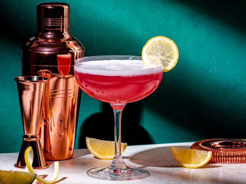 Veganer Pink Lady Cocktail in Glas mit Zitronenschreib, daneben Shaker und Zitronenspalten.