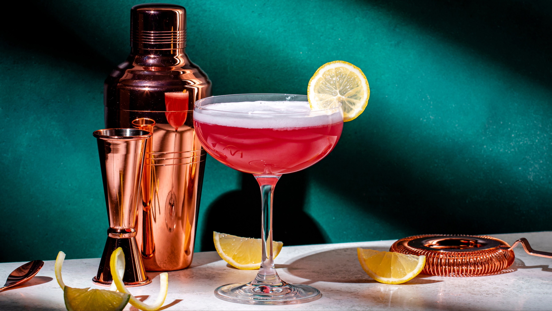 Veganer Pink Lady Cocktail in Glas mit Zitronenschreib, daneben Shaker und Zitronenspalten.