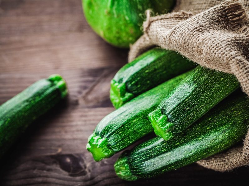 Frische grüne Zucchini liegen teilweise in einem groben Jutesack auf einer rustikalen Holzoberfläche.