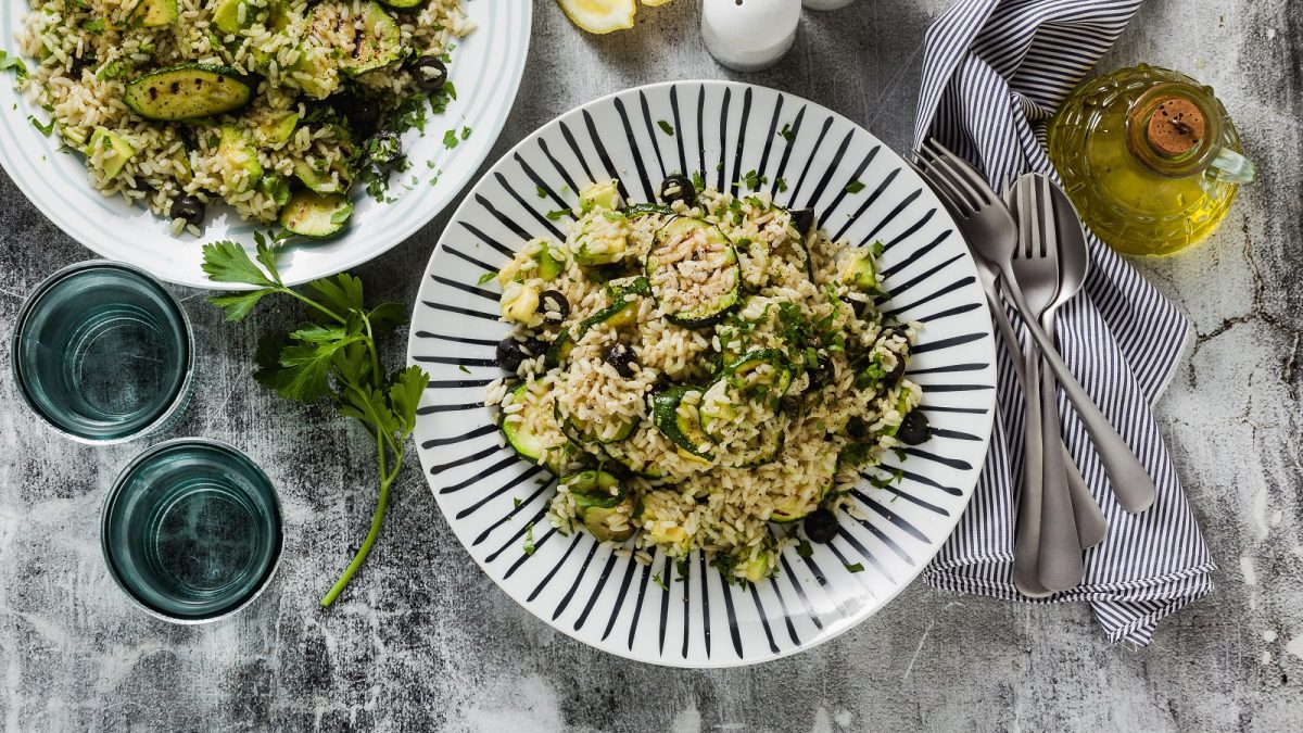 Zwei helle Teller mit der Zucchini-Pfanne auf einem grauen Steintisch, mit Gläsern, blau-weiß-gestreiftem Tuch sowie Olivenöl und Salz- und Pfefferstreuer. Alles direkt von oben fotografiert.