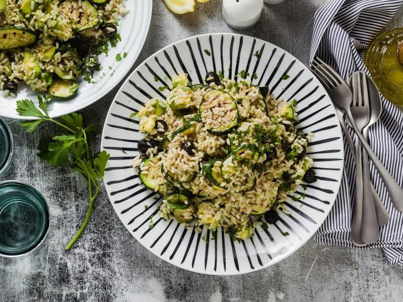 Zwei helle Teller mit der Zucchini-Pfanne auf einem grauen Steintisch, mit Gläsern, blau-weiß-gestreiftem Tuch sowie Olivenöl und Salz- und Pfefferstreuer. Alles direkt von oben fotografiert.