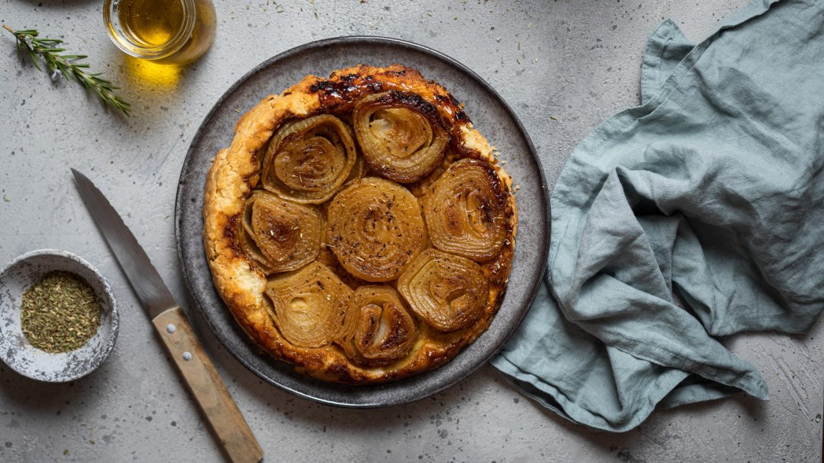 Draufsicht: Zwiebel-Tarte-Tatin auf Teller, daneben Messer, Küchentuch, Gewürze und Öl.