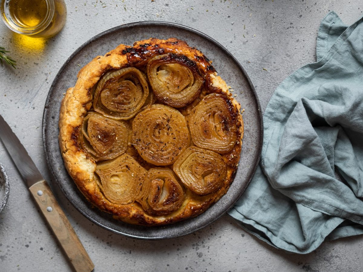 Draufsicht: Zwiebel-Tarte-Tatin auf Teller, daneben Messer, Küchentuch, Gewürze und Öl.