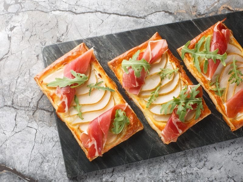 Drei Blätterteig-Snacks mit Birnenscheiben, Parmaschinken und Rucola von oben fotografiert auf einer schwarzen Steinplatte vor Marmor-Untergrund.