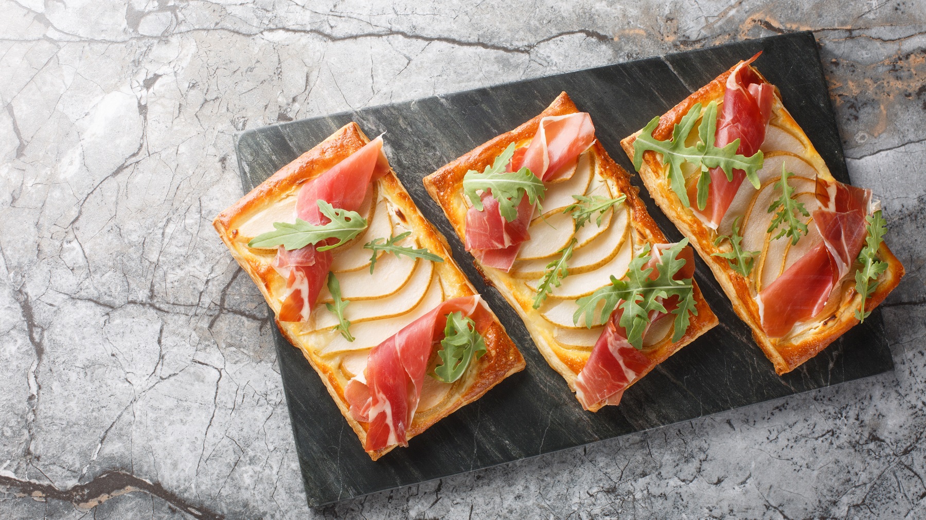 Drei Blätterteig-Snacks mit Birnenscheiben, Parmaschinken und Rucola von oben fotografiert auf einer schwarzen Steinplatte vor Marmor-Untergrund.