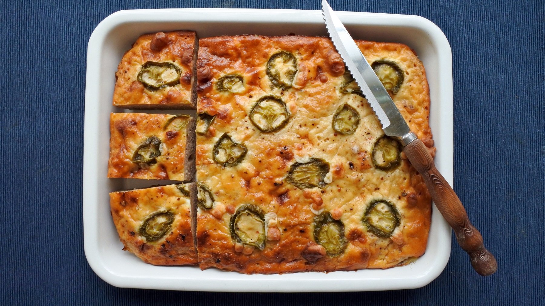 Eine Ofenform mit dem Maisbrot direkt von oben auf dunkelblauem, mattem Untergrund fotografiert. An der Seite liegt noch ein Messer mit Holzgriff.