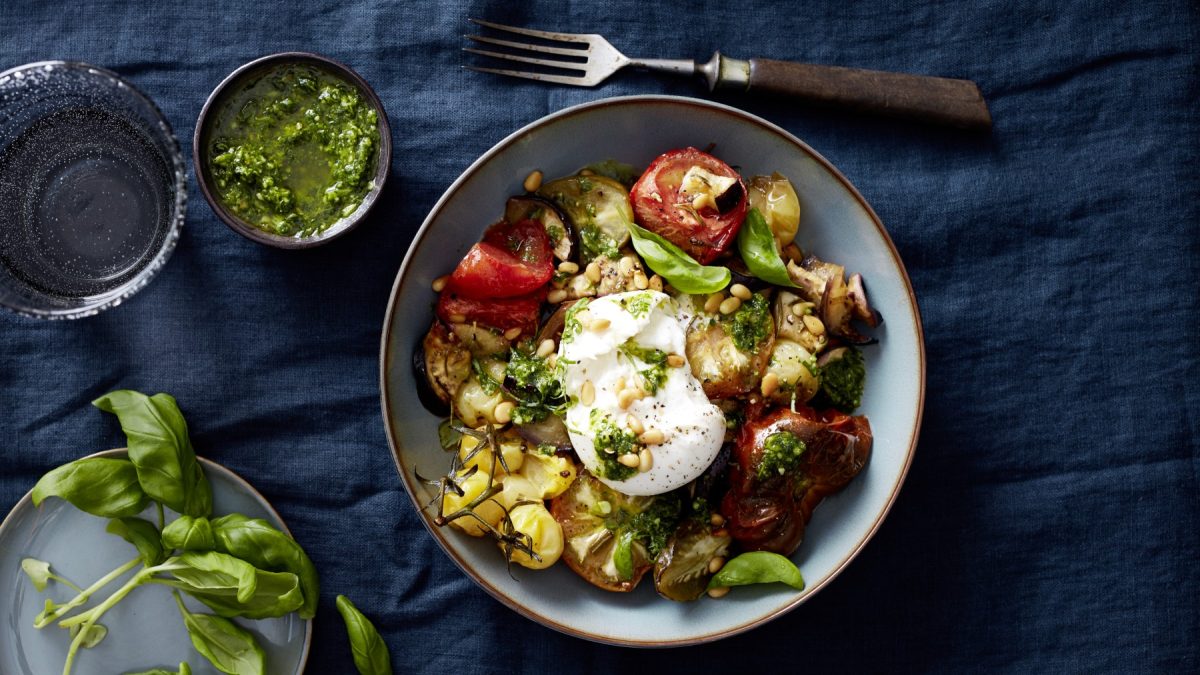 Draufsicht: Burratabowl mit Ofentomaten in einer Schüssel und daneben eine kleinere Schüssel mit Pesto und Deko auf dunkelblauem Stoffuntergrund.