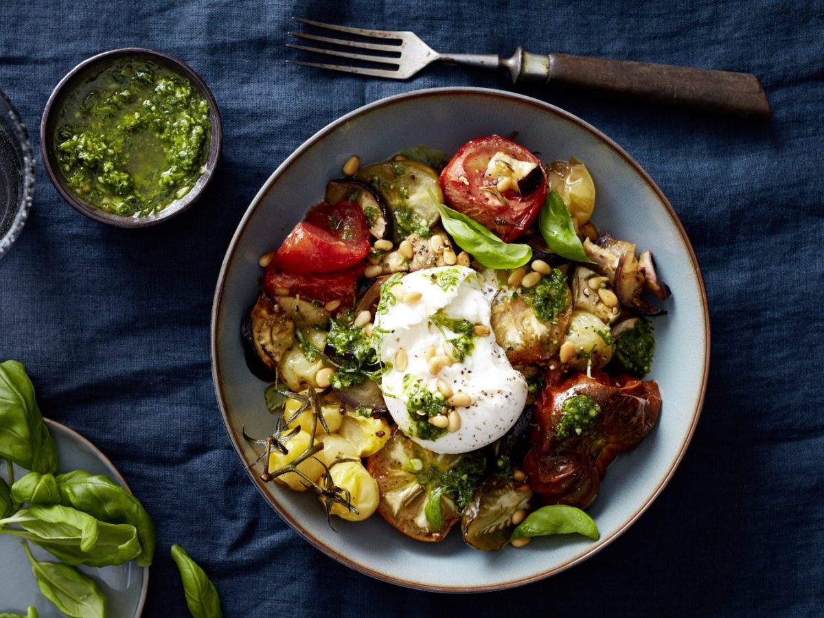 Draufsicht: Burratabowl mit Ofentomaten in einer Schüssel und daneben eine kleinere Schüssel mit Pesto und Deko auf dunkelblauem Stoffuntergrund.