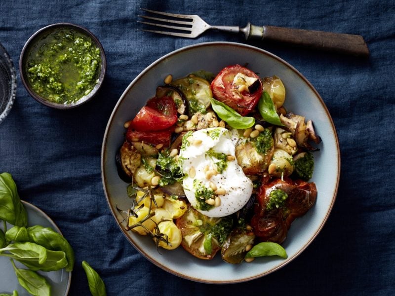 Draufsicht: Burratabowl mit Ofentomaten in einer Schüssel und daneben eine kleinere Schüssel mit Pesto und Deko auf dunkelblauem Stoffuntergrund.