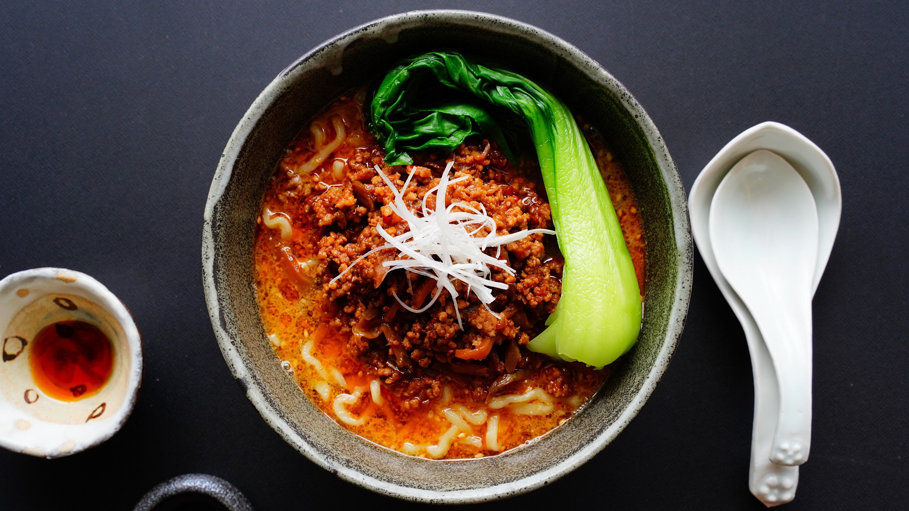 Eine graue Schale mit dem chinesischen Nudelgericht auf dunkelblauem Untergrund direkt von oben fotografiert. Rechts daneben asiatische Suppenlöffel und links daneben ein Schälchen mit Chiliöl.