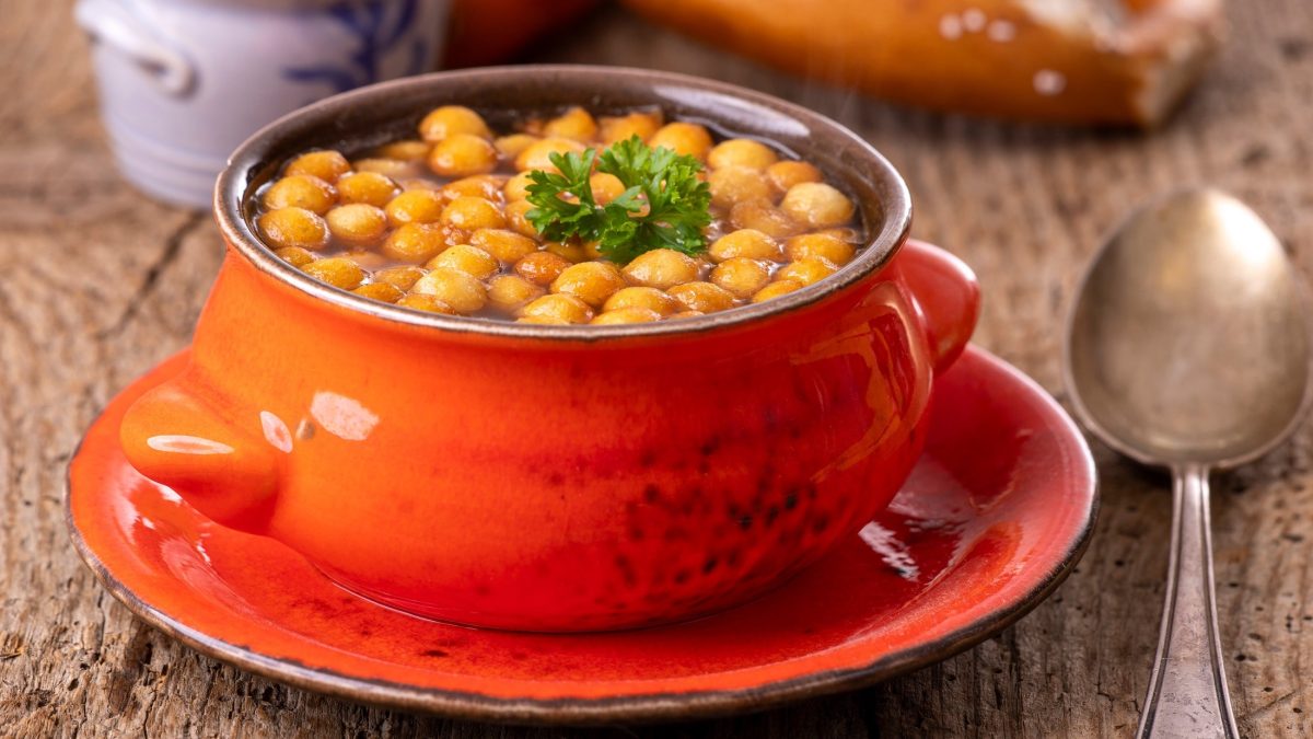 Frontal: Eine orangefarbene Suppentasse mit einer Suppe und selbst gemachten Backerbsen auf einer gleichfarbigen Untertasse. Die Tasse steht auf einem Untergrund aus Holz. Im Hintergrund ist eine bayerische Breze. / Backerbsen selbst machen