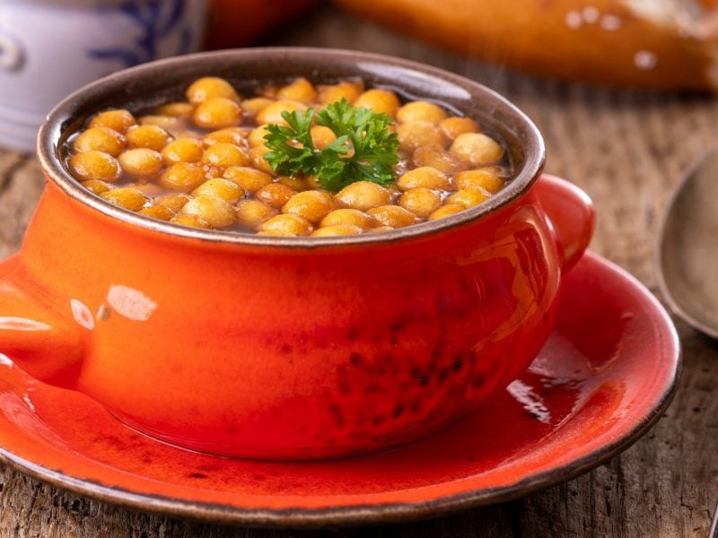 Frontal: Eine orangefarbene Suppentasse mit einer Suppe und selbst gemachten Backerbsen auf einer gleichfarbigen Untertasse. Die Tasse steht auf einem Untergrund aus Holz. Im Hintergrund ist eine bayerische Breze. / Backerbsen selbst machen