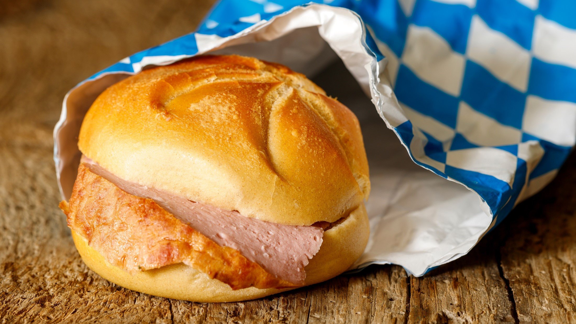 Eine Leberkässemmel mit selbst gebackenem bayerischen Leberkäse liegt halb in einer Papiertüte, die mit der bayerischen Flagge bedruckt ist auf einem Untergrund aus Holz.