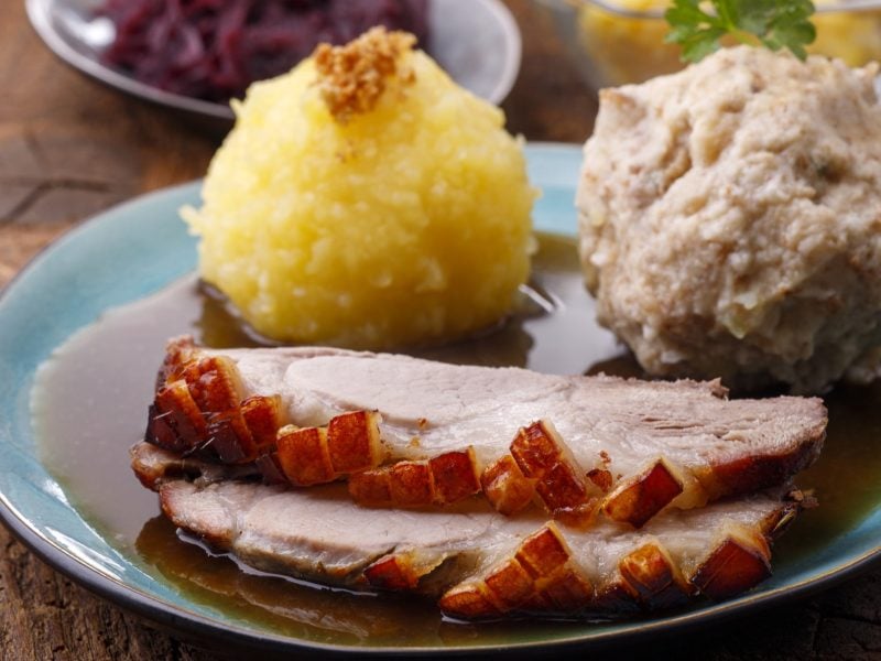 Draufsicht schräg: Eine Teller Bayerischer Schweinebraten in Biersauce sowie zwei unterschiedlichen Klößen auf einem rustikalen Untergrund aus Holz.