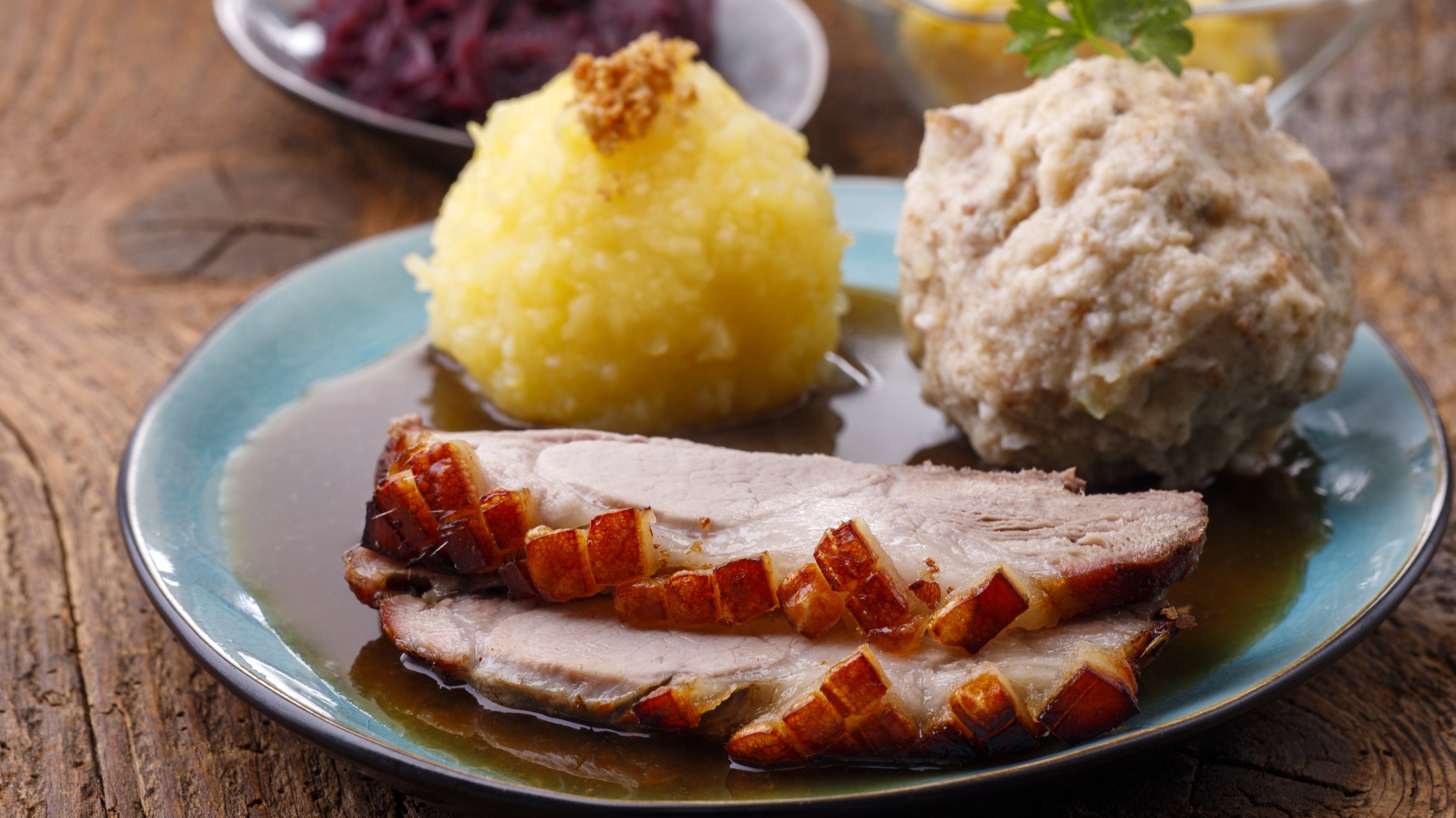 Draufsicht schräg: Eine Teller Bayerischer Schweinebraten in Biersauce sowie zwei unterschiedlichen Klößen auf einem rustikalen Untergrund aus Holz.