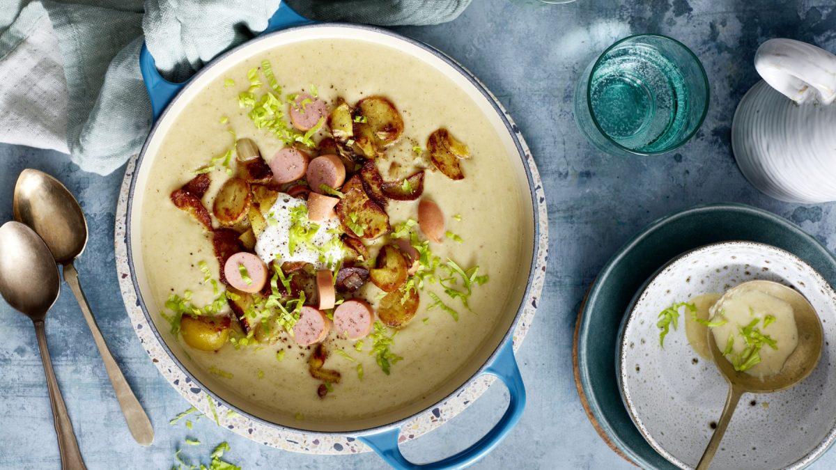 Bratkartoffel-Suppe mit Würstchen in einem Topf auf grauem Hintergrund. Links ein Löffel, rechts Wasser und eine Schöpfkelle.