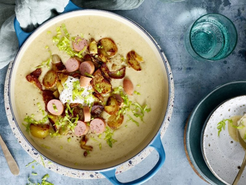 Bratkartoffel-Suppe mit Würstchen in einem Topf auf grauem Hintergrund. Links ein Löffel, rechts Wasser und eine Schöpfkelle.