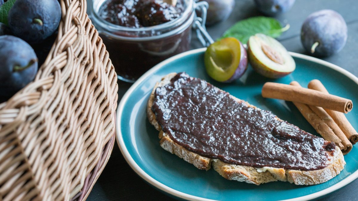 Draufsicht schräg: Ein Glas selbst gemachten Brotaufstrich mit Zwetschge und Zimt. Daneben lieget ein Brot, dass mit der Marmelade bestrichen ist. Und da neben liegen frische Zwetchgen sowie ein Korb mit dem Steinobst.