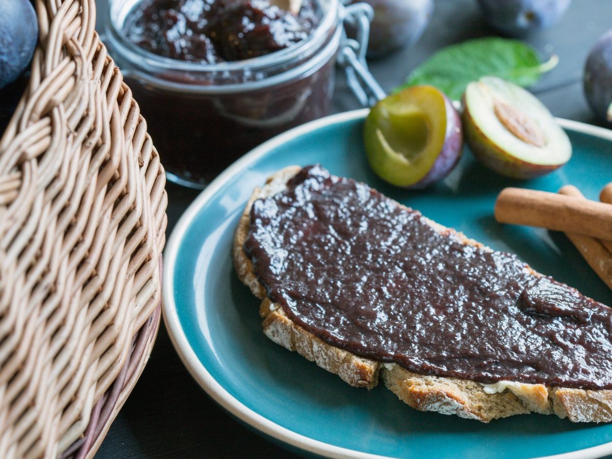 Draufsicht schräg: Ein Glas selbst gemachten Brotaufstrich mit Zwetschge und Zimt. Daneben lieget ein Brot, dass mit der Marmelade bestrichen ist. Und da neben liegen frische Zwetchgen sowie ein Korb mit dem Steinobst.