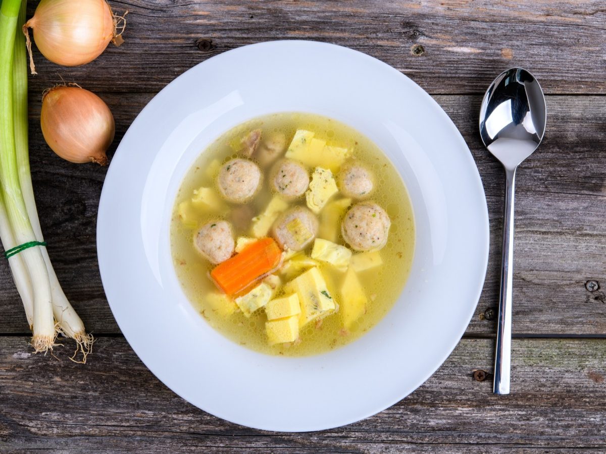 Draufsicht: Ein weißer teller mit einer klaren Suppe. In der sind Fleischbällchen, Möhren und selbst gemachter Eierstich. Daneben liegen Lauchzwiebeln, zwei Eier und ein Suppenlöffel. Der Untergrund ist aus Holz.