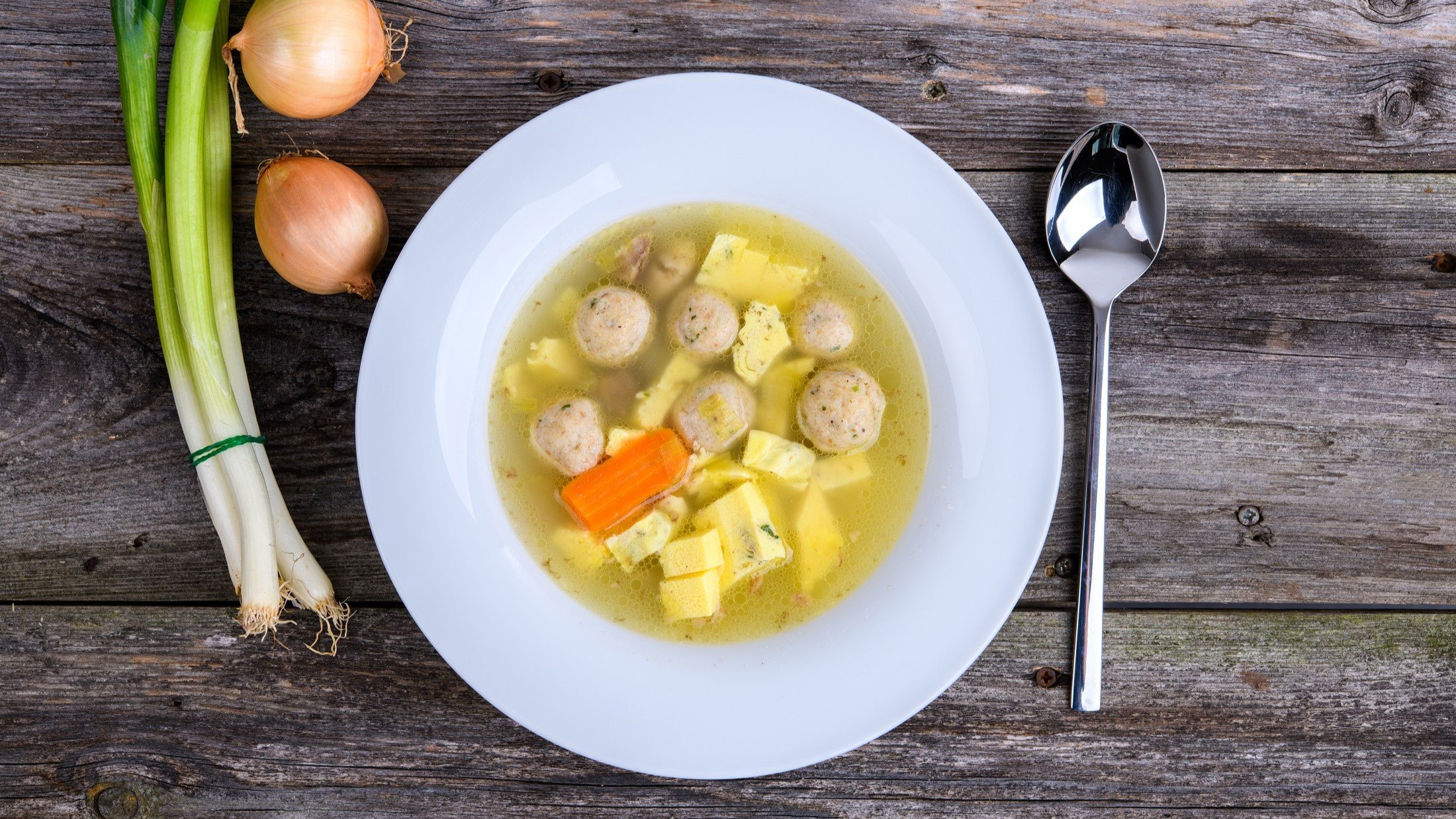 Draufsicht: Ein weißer teller mit einer klaren Suppe. In der sind Fleischbällchen, Möhren und selbst gemachter Eierstich. Daneben liegen Lauchzwiebeln, zwei Eier und ein Suppenlöffel. Der Untergrund ist aus Holz.