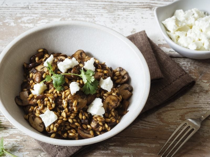 Draufblick, schräg: Ein weiße Schüssel Gersten-Risotto mit Feta und Pilzen steht auf einem Holzboden. Daneben ist eine kleinere weiße Schüssel mit gewürfeltem Feta.