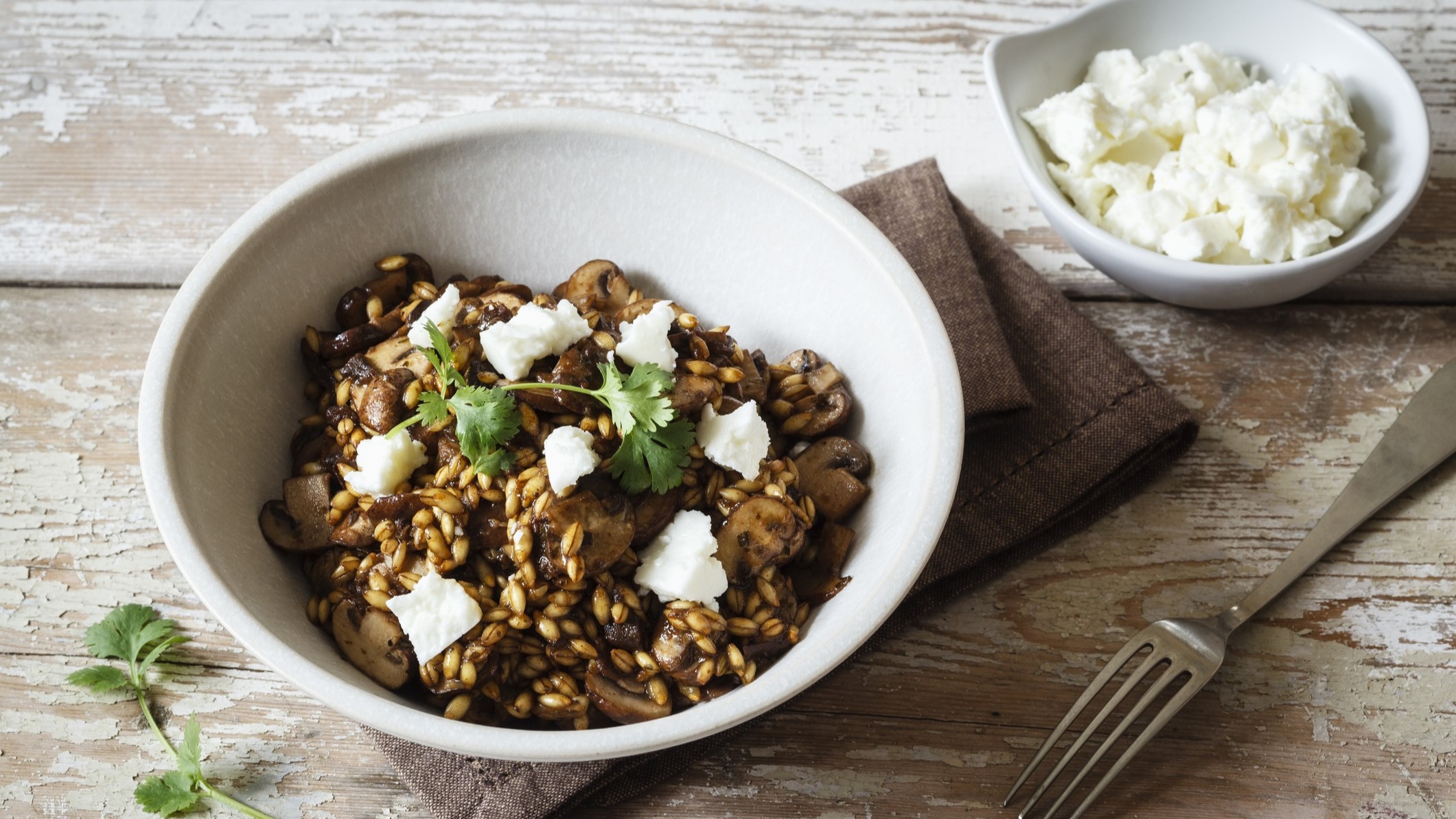 Draufblick, schräg: Ein weiße Schüssel Gersten-Risotto mit Feta und Pilzen steht auf einem Holzboden. Daneben ist eine kleinere weiße Schüssel mit gewürfeltem Feta.