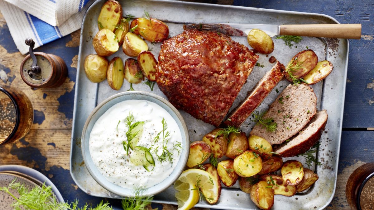 Glasierter Hackbraten auf einem silbernem Blech auf hölzernem Hintergrund. Links auf dem Foto eine Pfeffermühle und ein Glas Wasser.