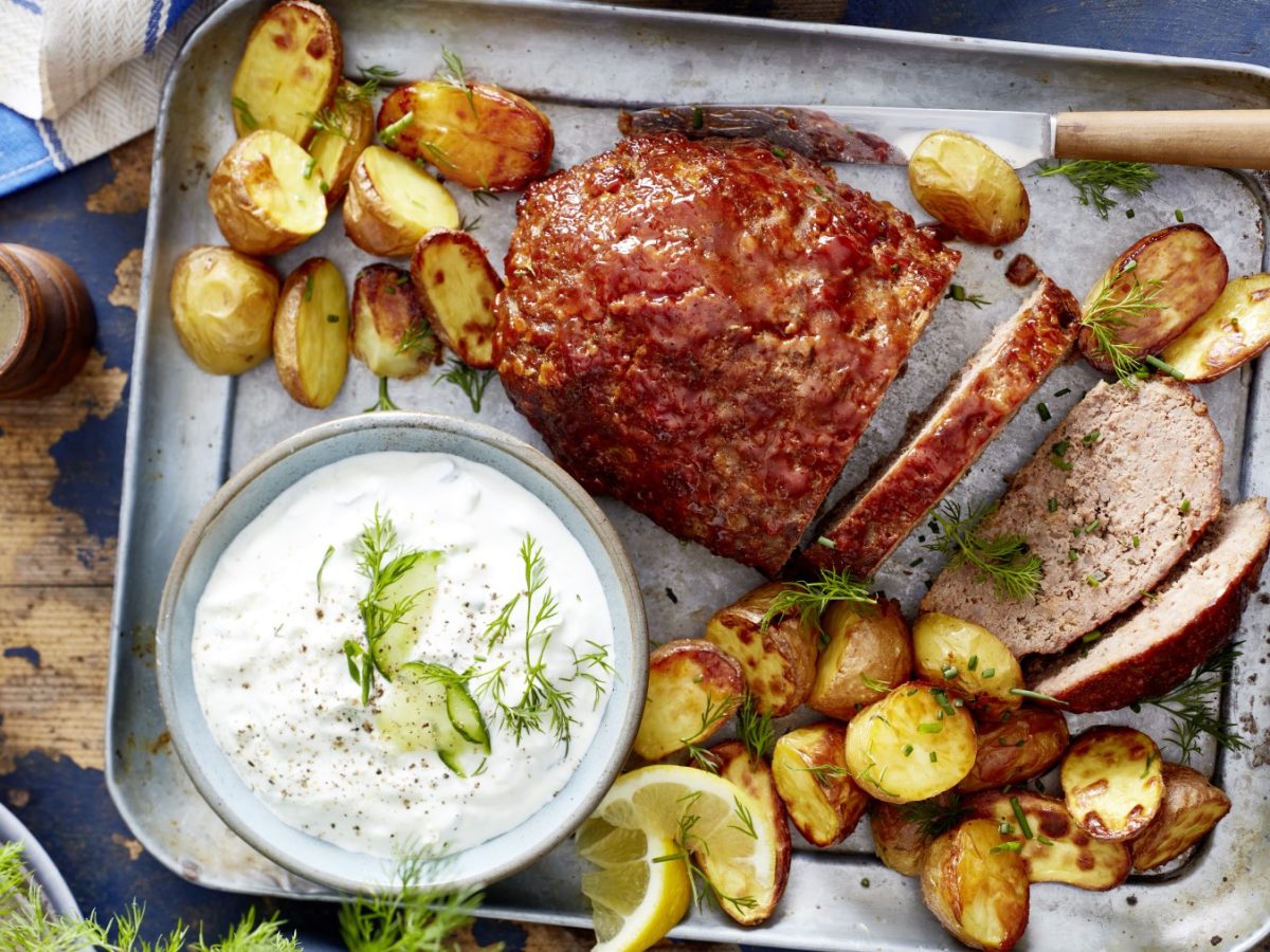 Glasierter Hackbraten auf einem silbernem Blech auf hölzernem Hintergrund. Links auf dem Foto eine Pfeffermühle und ein Glas Wasser.