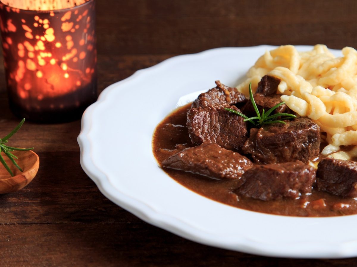 Draufsicht, schräg: Ein Hirschgulasch mit Spätzle auf einem weißen Essteller, der auf einem dunklen Holztisch steht.