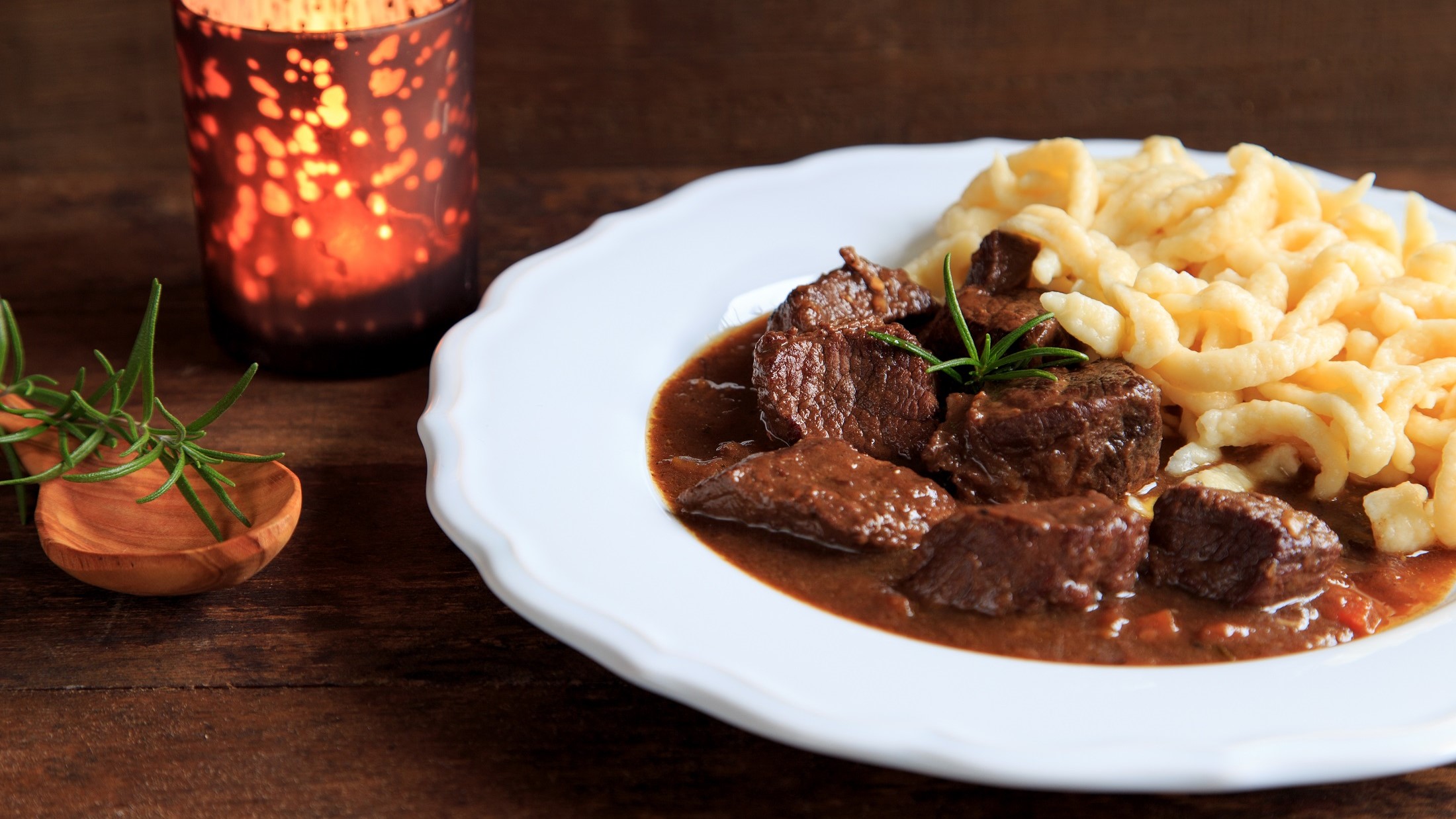 Draufsicht, schräg: Ein Hirschgulasch mit Spätzle auf einem weißen Essteller, der auf einem dunklen Holztisch steht.