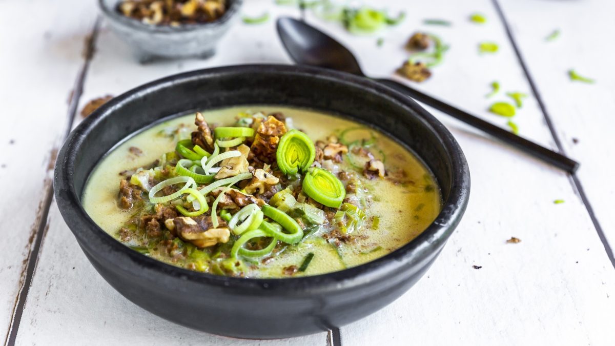 Draufsicht, schräg: Eine schwarze Schüssel Käse-Lauch-Suppe mit Walnüssen auf einem weißen Untergrund aus Holzdielen. Daneben liegen ein Suppenlöffel, lauchringe und eine Schale mit Walnusskernen.